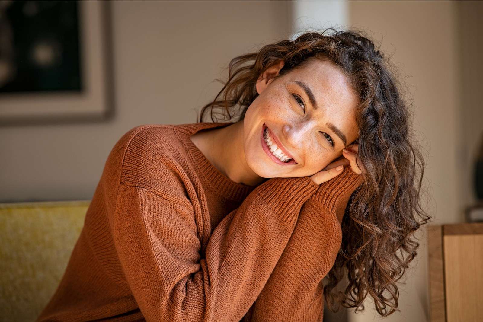 A woman smiling warmly, resting her chin on her hand in soft, warm light.