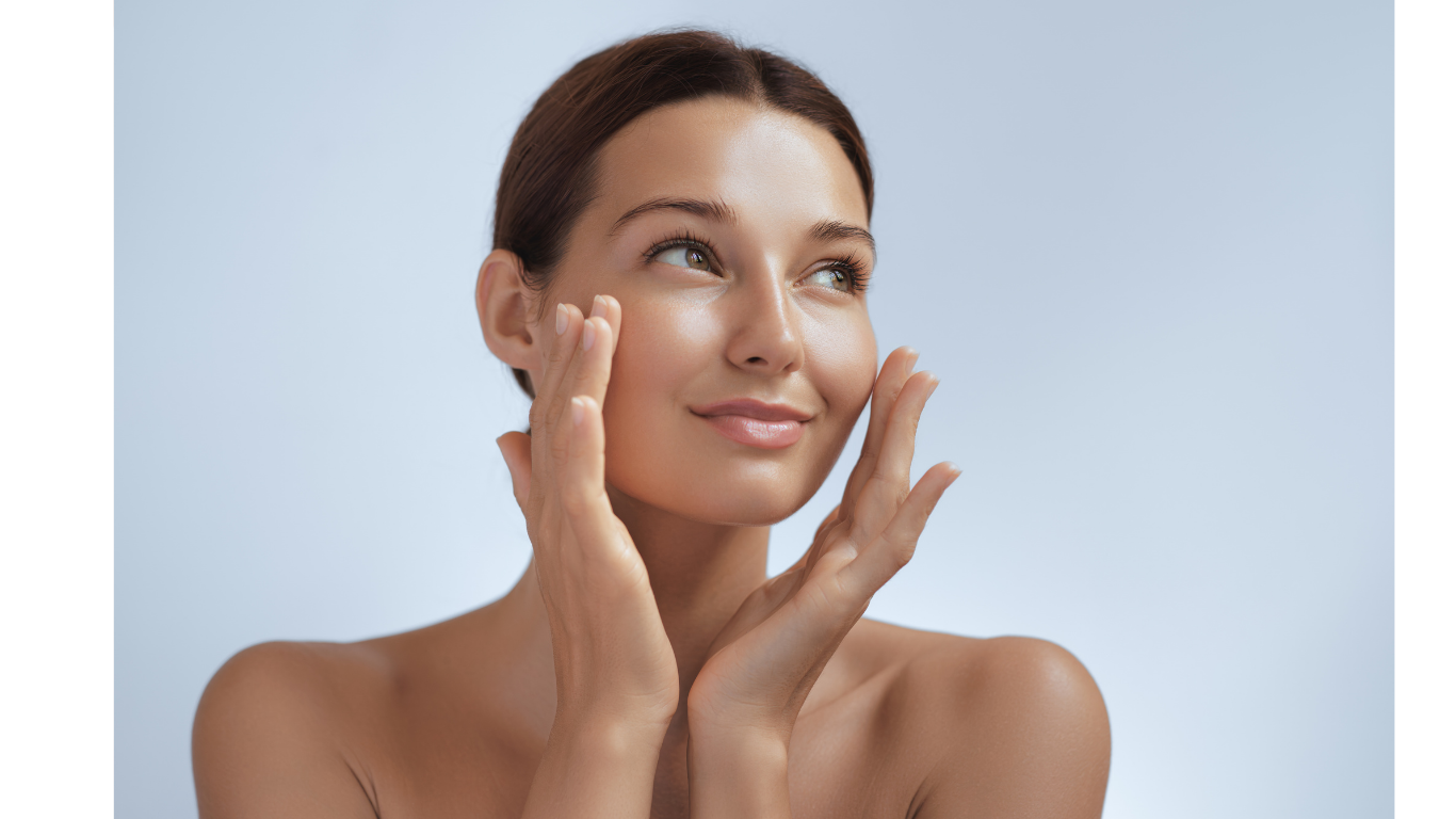 A woman with radiant skin framing her face with her hands in soft light.